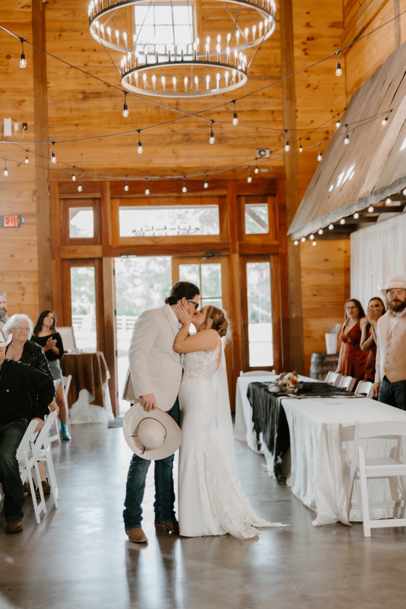 Shay & Wyatt kiss in the Covey Creek reception barn beneath circular chandeliers and draped string lights.
