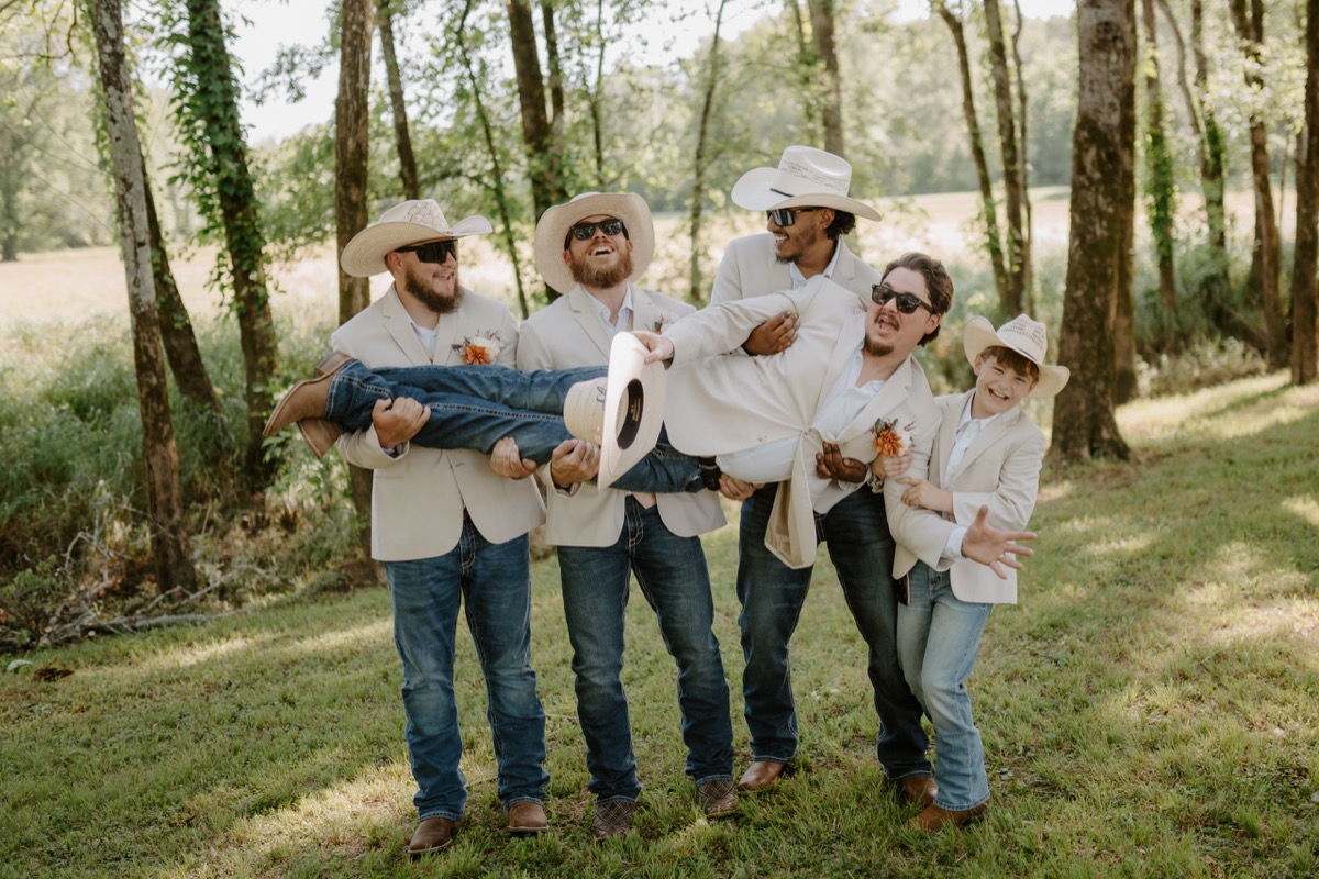 Groomsmen in cowboy hats lift Wyatt overhead in a sunny field at Covey Creek for Shay & Wyatt's wedding.