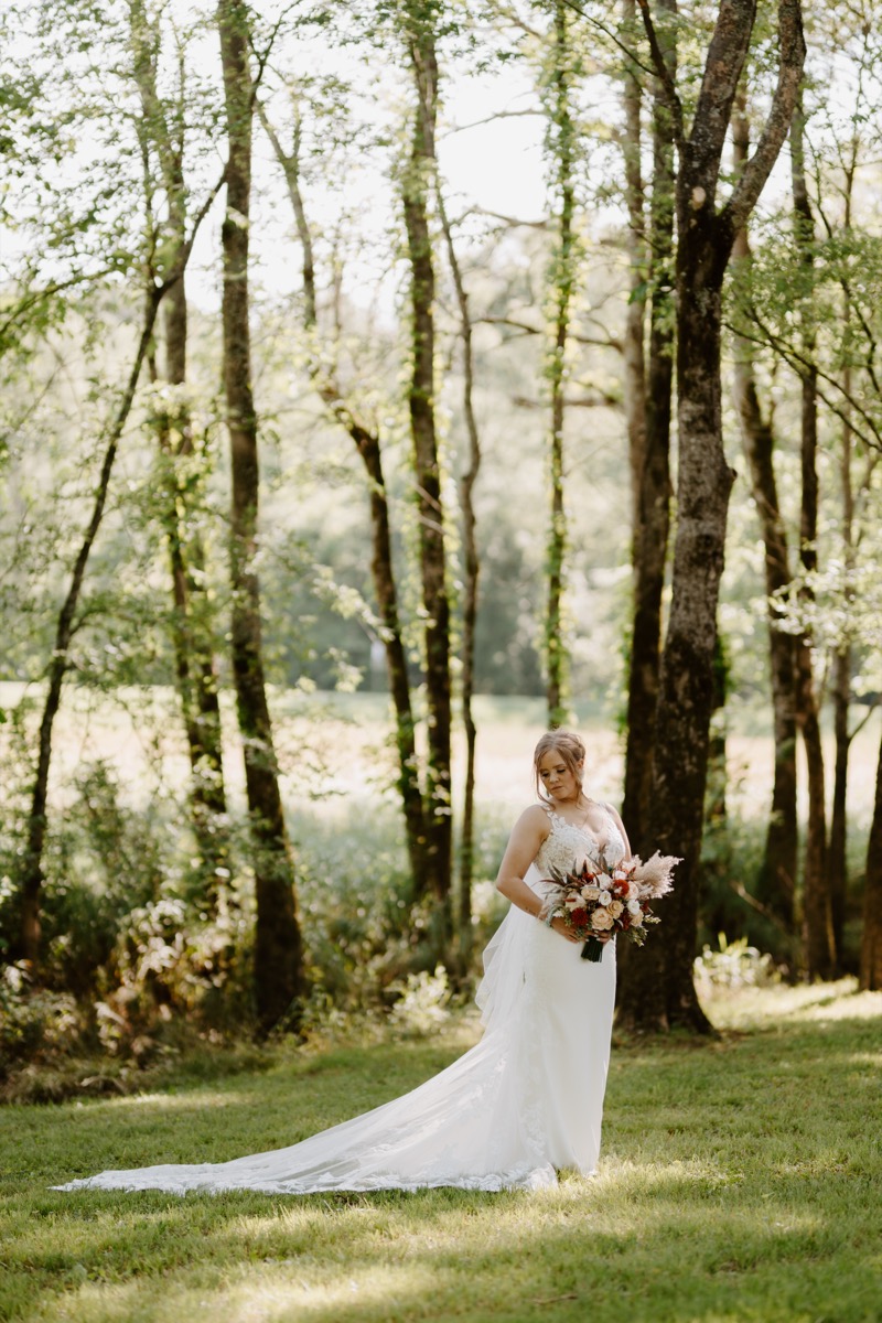 Shay stands among sunlit trees at Covey Creek for Shay & Wyatt's wedding, orange bouquet and lace train shown.