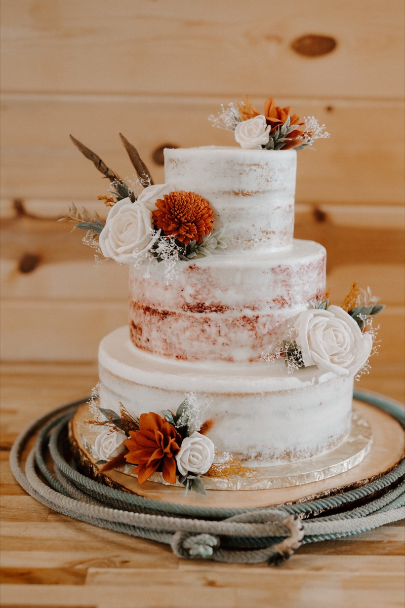 Shay & Wyatt's three-tier semi-naked cake with white roses and orange blooms on a wood slice at Covey Creek.