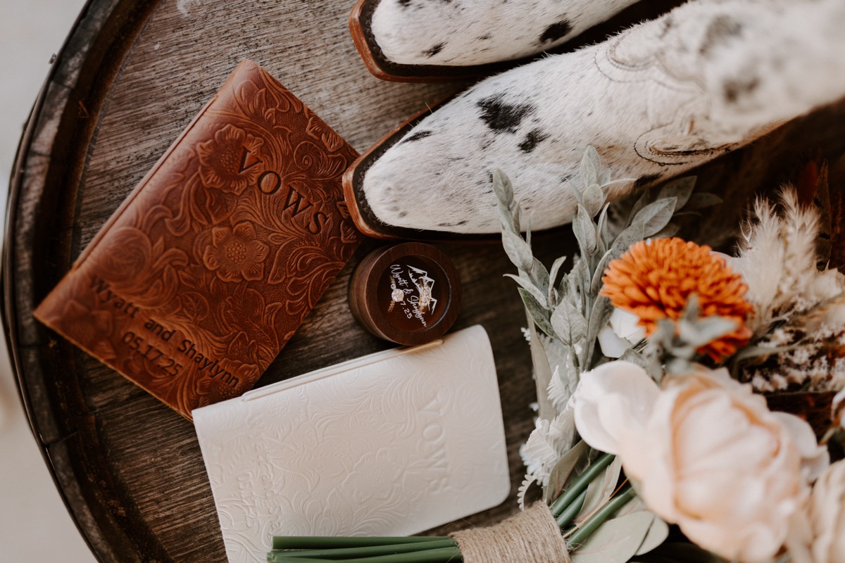 Shay & Wyatt's wedding details at Covey Creek: leather vows books, ring box, cowhide boots, and orange bouquet.