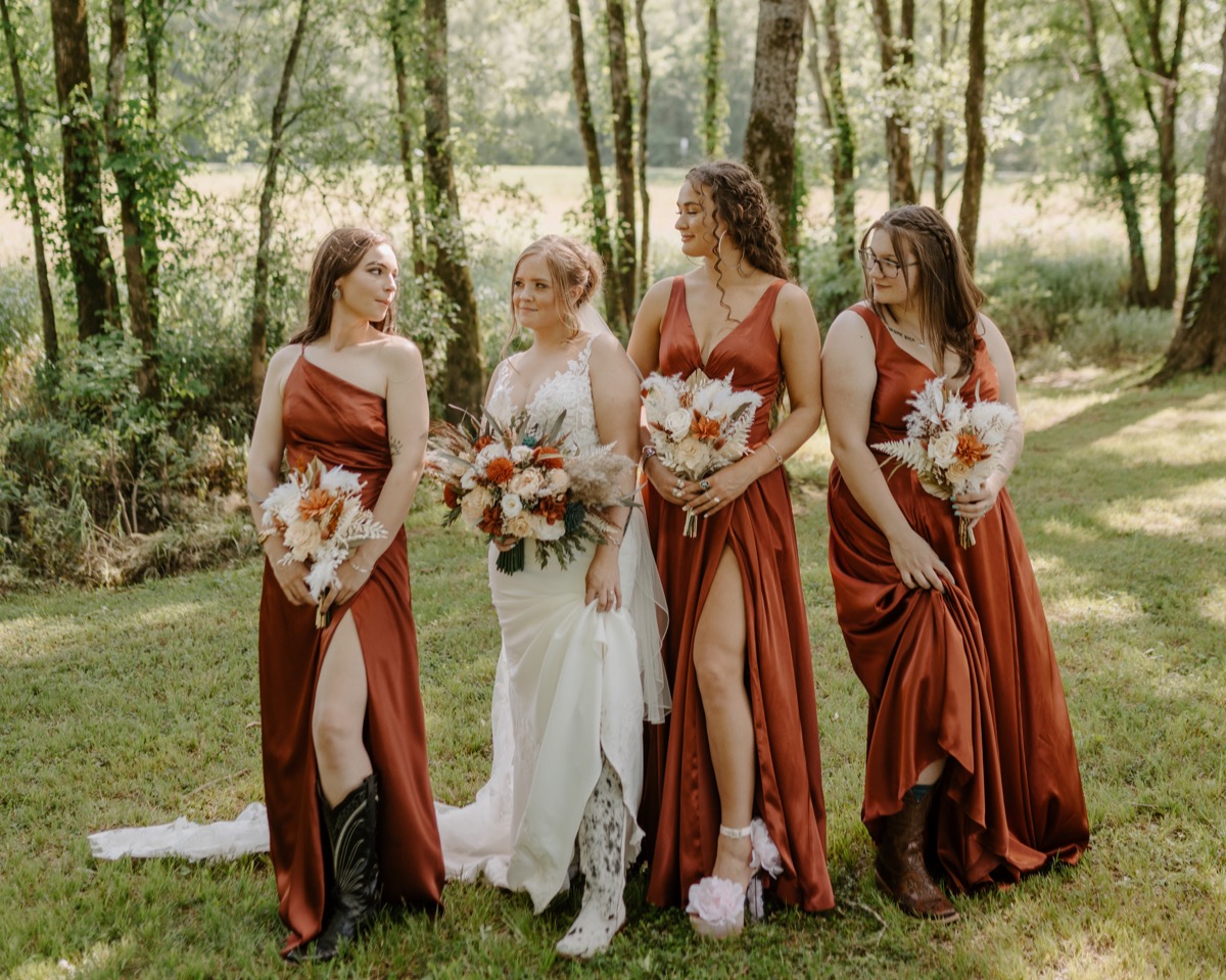Shay & Wyatt's bridal party lifts rust dresses to show cowboy boots and heels in a grassy clearing at Covey Creek.