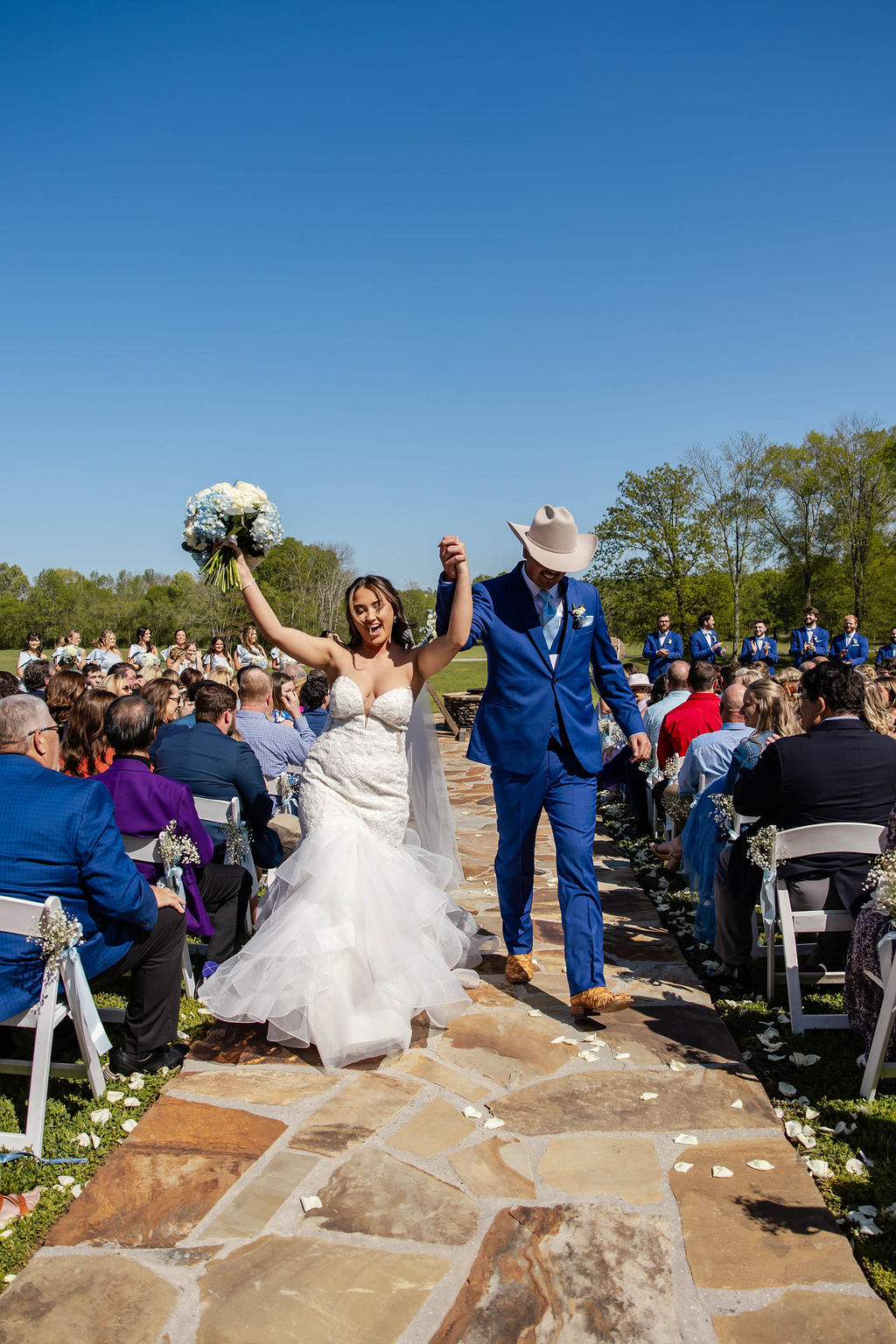 Shelbie & Callaway cheer down the Firepit stone aisle, bouquet raised, blue suit and cowboy hat at Covey Creek