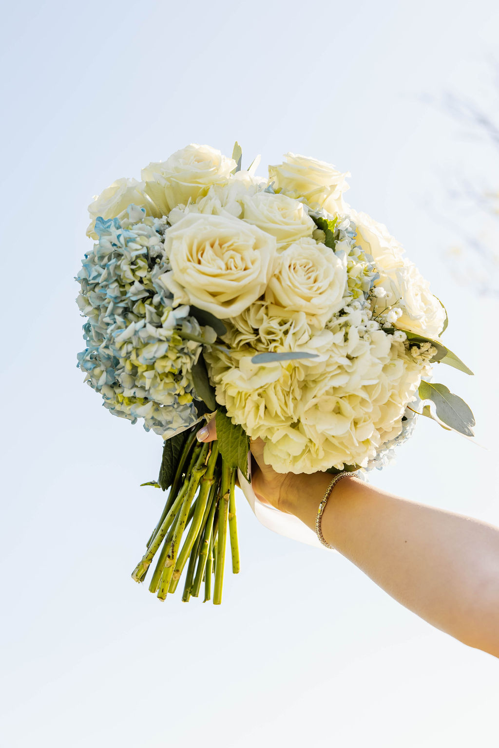 Shelbie & Callaway bridal bouquet of white roses and blue hydrangeas held high against a clear Covey Creek sky