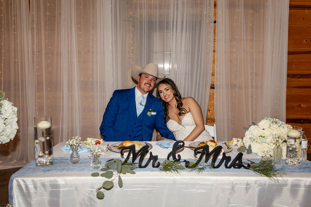 Shelbie & Callaway smile at their Mr & Mrs sweetheart table with blue florals and fairy lights at Covey Creek