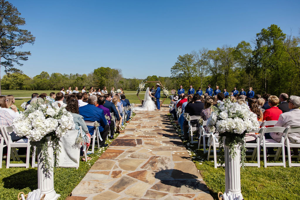 Shelbie & Callaway stand under a floral hex arbor as guests fill white chairs at Covey Creek Firepit ceremony