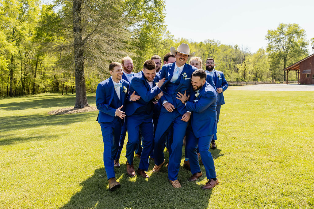 Shelbie & Callaway: groomsmen in blue suits lift Callaway in a cowboy hat on the lawn at Covey Creek