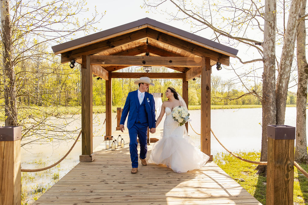 Shelbie & Callaway walk hand in hand on the Pond pavilion dock with lanterns and water behind at Covey Creek