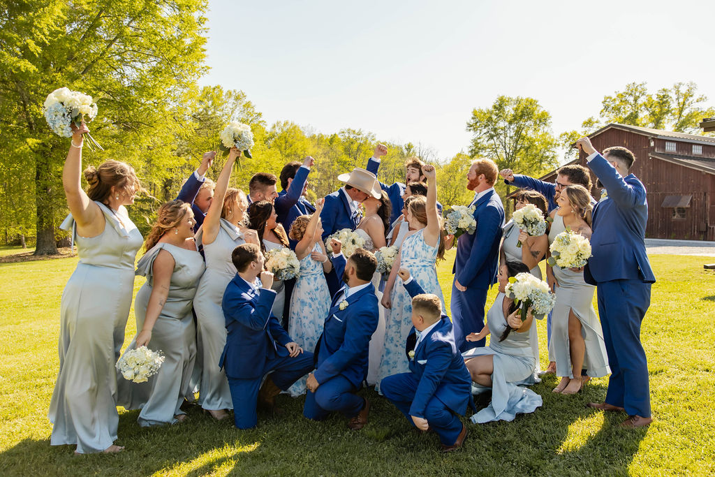 Shelbie & Callaway kiss as their blue wedding party cheers on the sunny lawn by the barn at Covey Creek