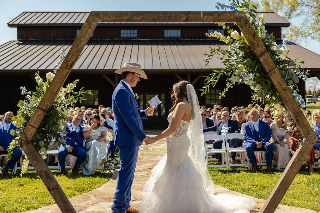 Shelbie & Callaway exchange vows under a floral hex arbor with barn and guests behind at Covey Creek Firepit