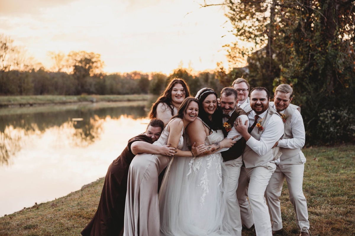 Tori & Tim laugh in a group hug with their wedding party beside the Pond at sunset, Covey Creek