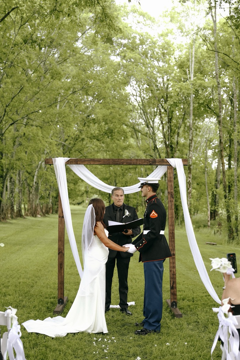 Cadence & Brody hold hands under a white-draped wood arch on the wooded lawn during vows at Covey Creek