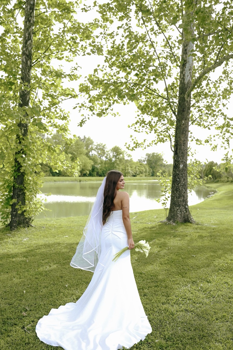 Cadence glances toward the Pond in gown and veil between trees at Cadence & Brody's Covey Creek wedding