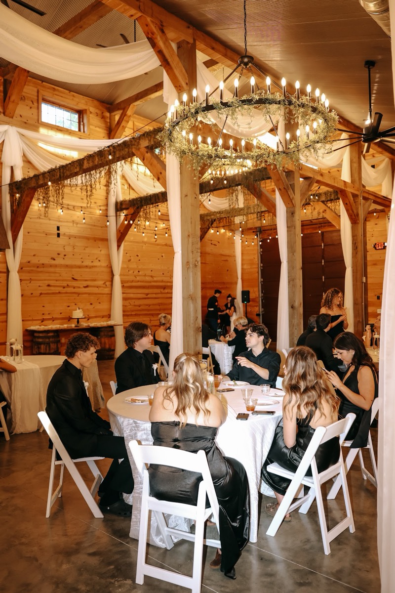 Round tables fill the barn under wagon-wheel chandeliers for Cadence & Brody's reception at Covey Creek