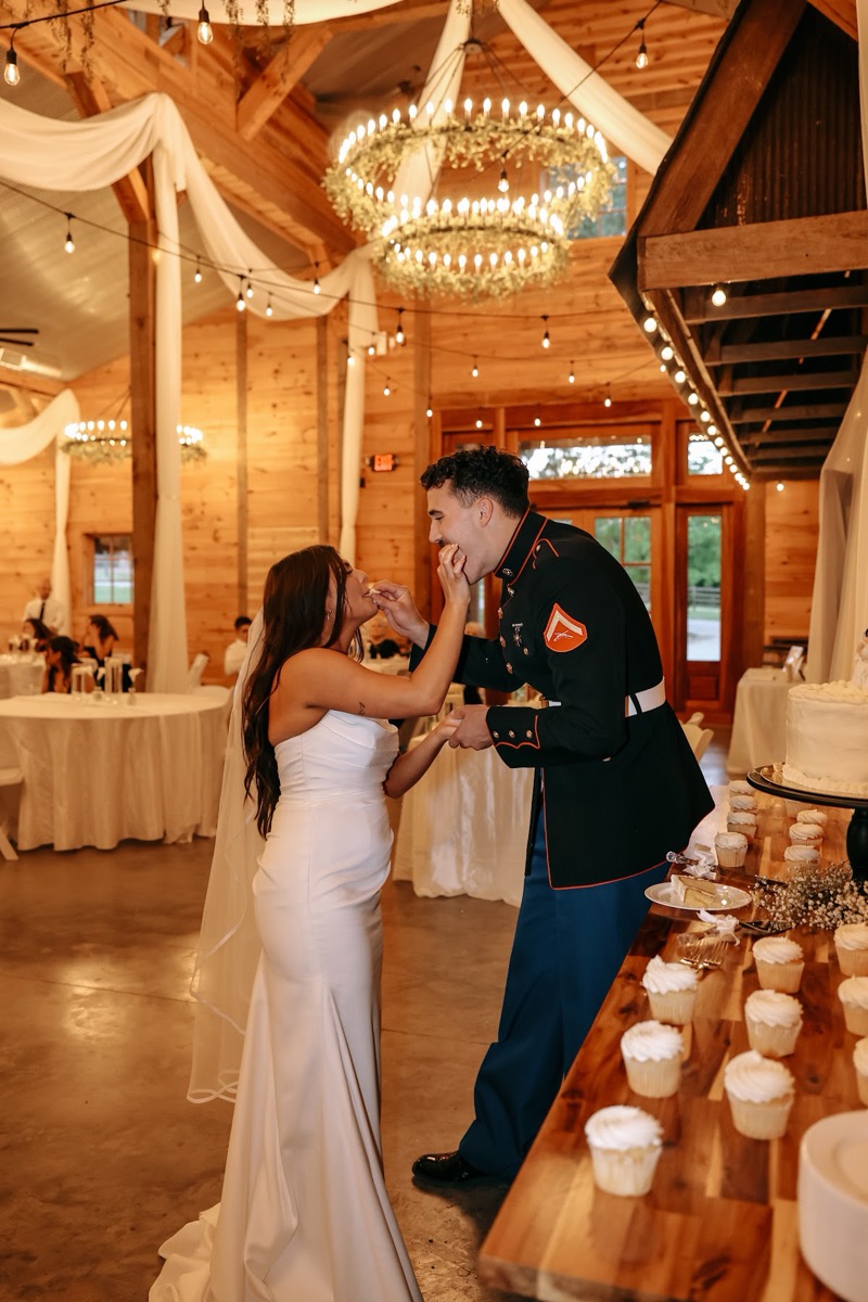 Cadence & Brody feed each other cake beside cupcakes under draped barn beams at Covey Creek