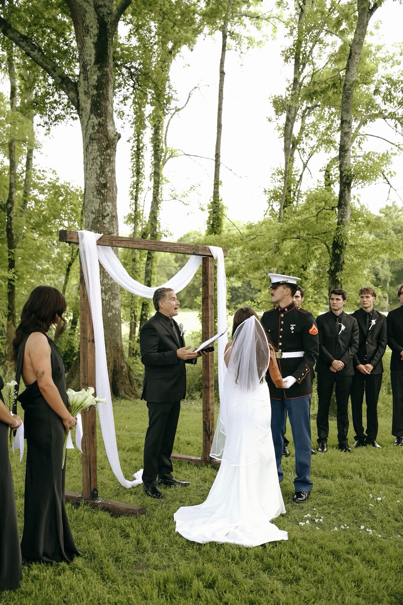 Cadence & Brody face the officiant under a white-draped wood arch with their party in black at Covey Creek