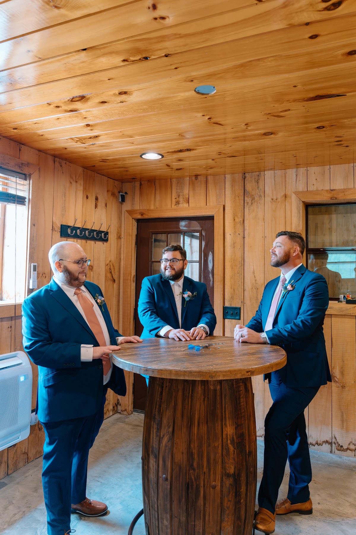 Justin and groomsmen in navy suits with peach ties chat around a barrel table indoors at Covey Creek for Katelynn & Justin