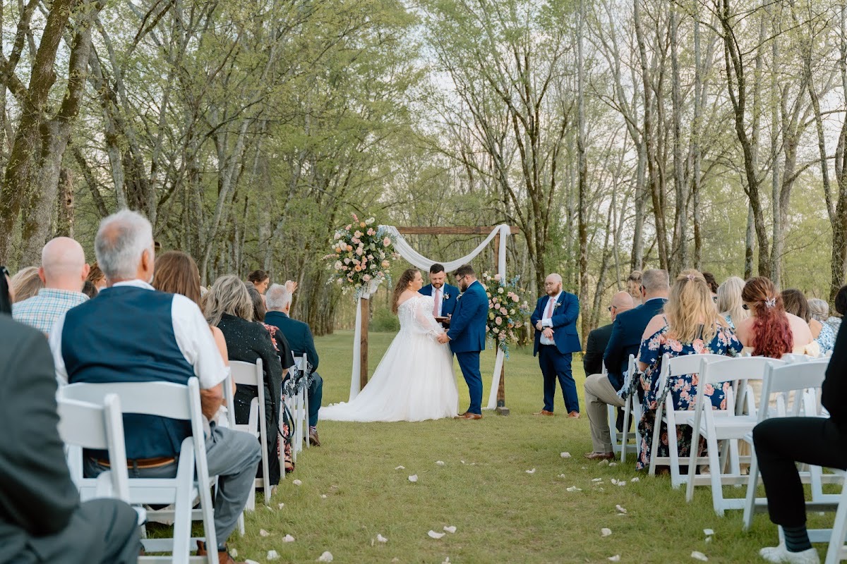 Katelynn & Justin stand at the floral arbor as guests fill white chairs for their outdoor ceremony at Covey Creek