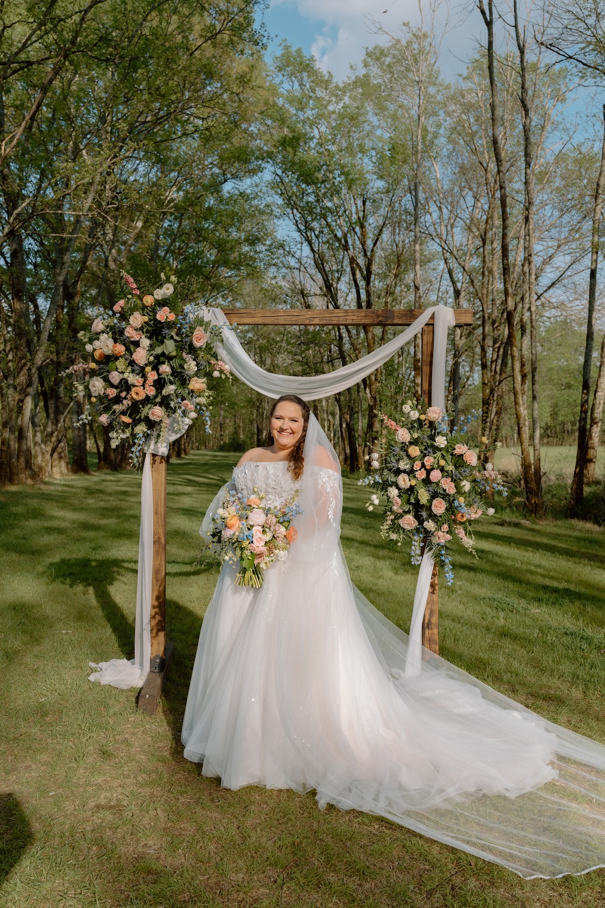 Katelynn stands alone under a floral wooden arbor in her lace gown and veil at Covey Creek for Katelynn & Justin