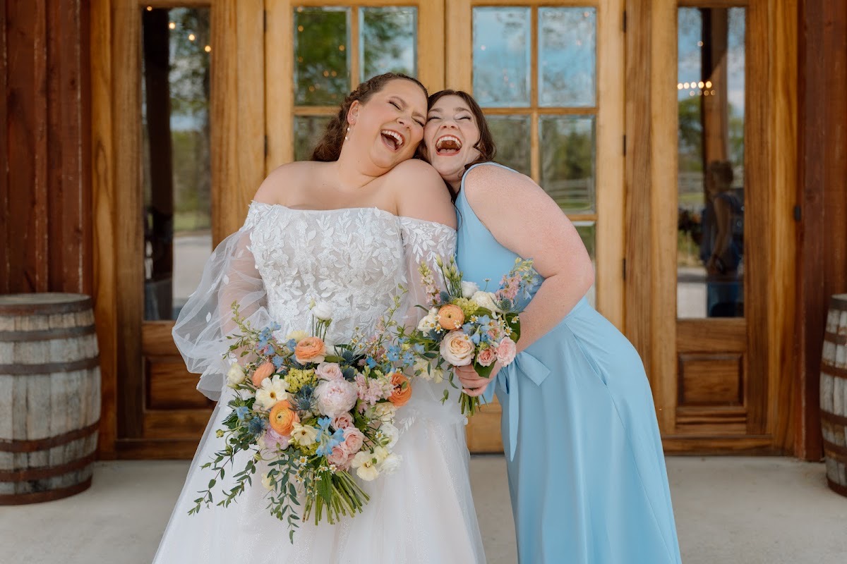 Katelynn laughs with a bridesmaid in blue, peach and blue bouquets raised by barn doors at Covey Creek for Katelynn & Justin