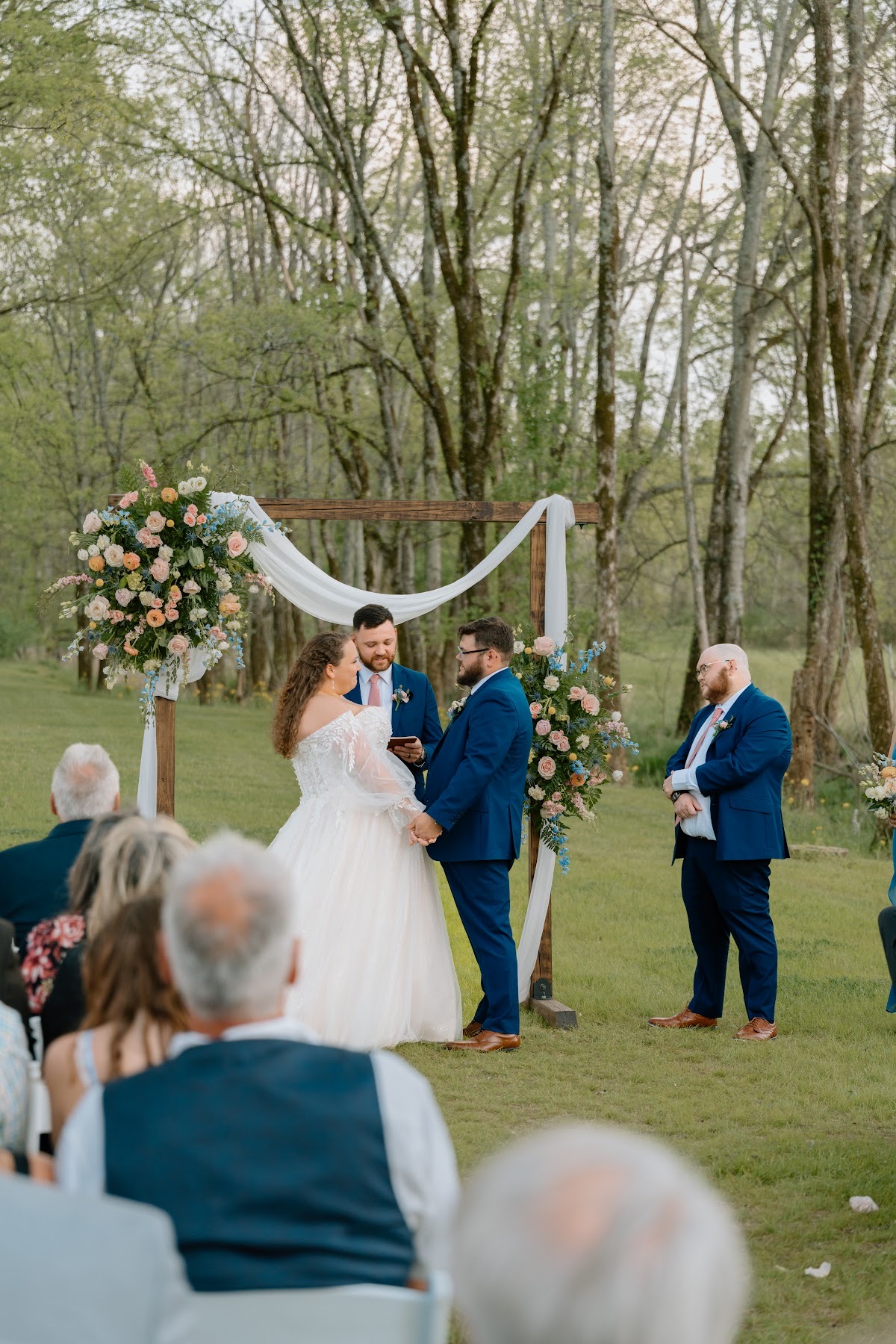 Katelynn & Justin hold hands under a floral wooden arbor during their outdoor ceremony among trees at Covey Creek