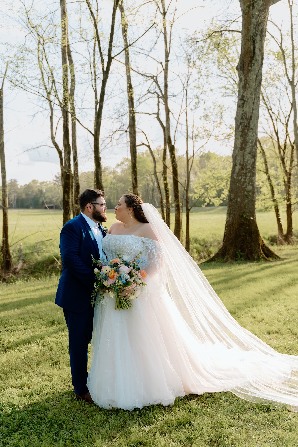 Katelynn & Justin lean in for a kiss on a sunny lawn, her veil blowing among trees at Covey Creek