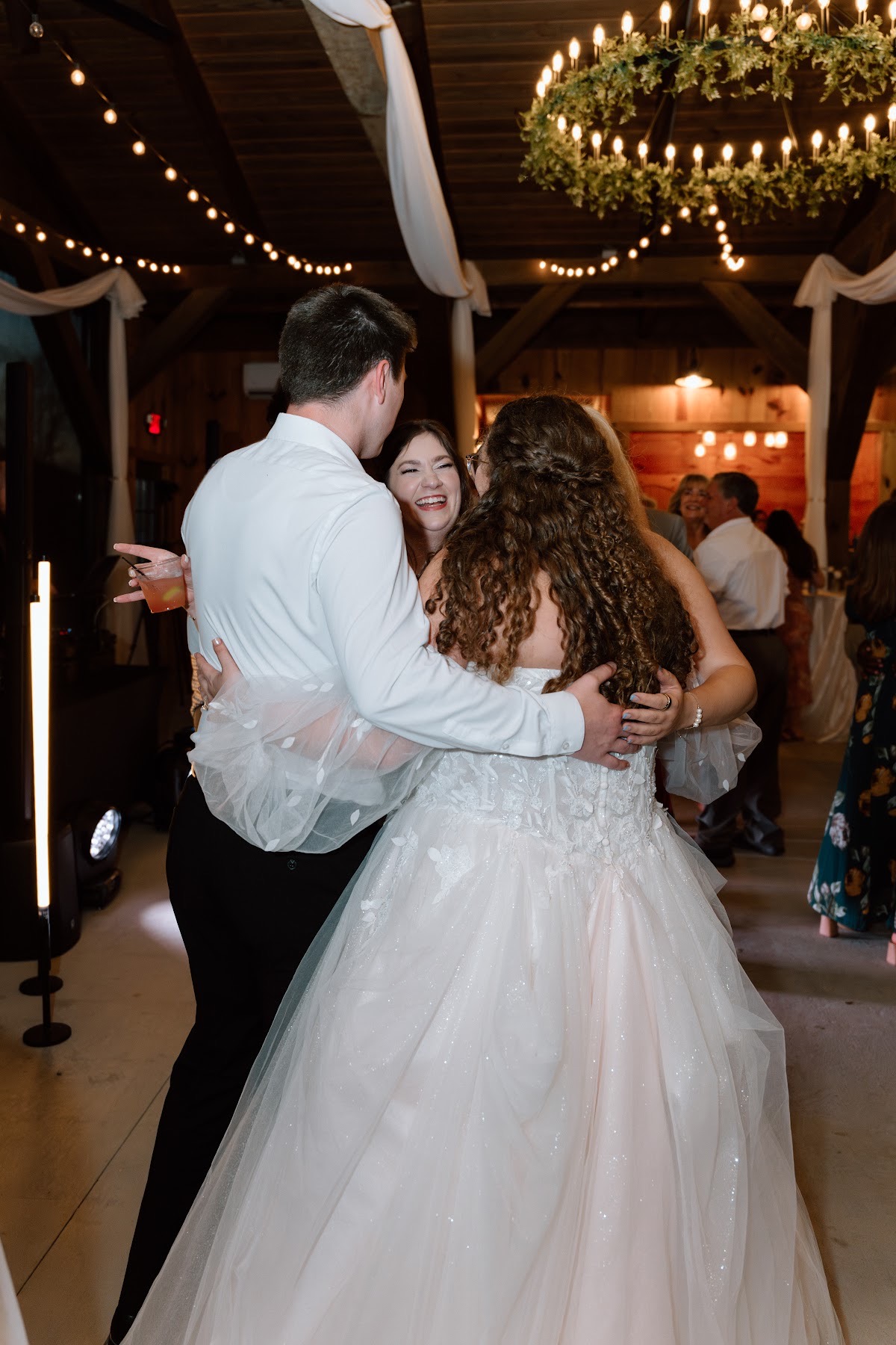 Katelynn & Justin hug a laughing guest under string lights and a barn chandelier at Covey Creek