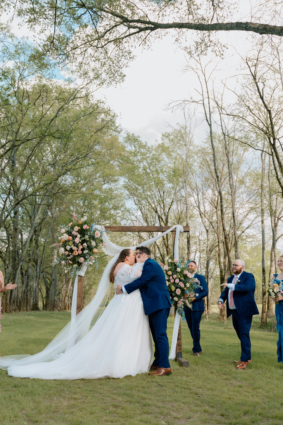 Katelynn & Justin share their first kiss under a floral wooden arbor as the wedding party cheers at Covey Creek