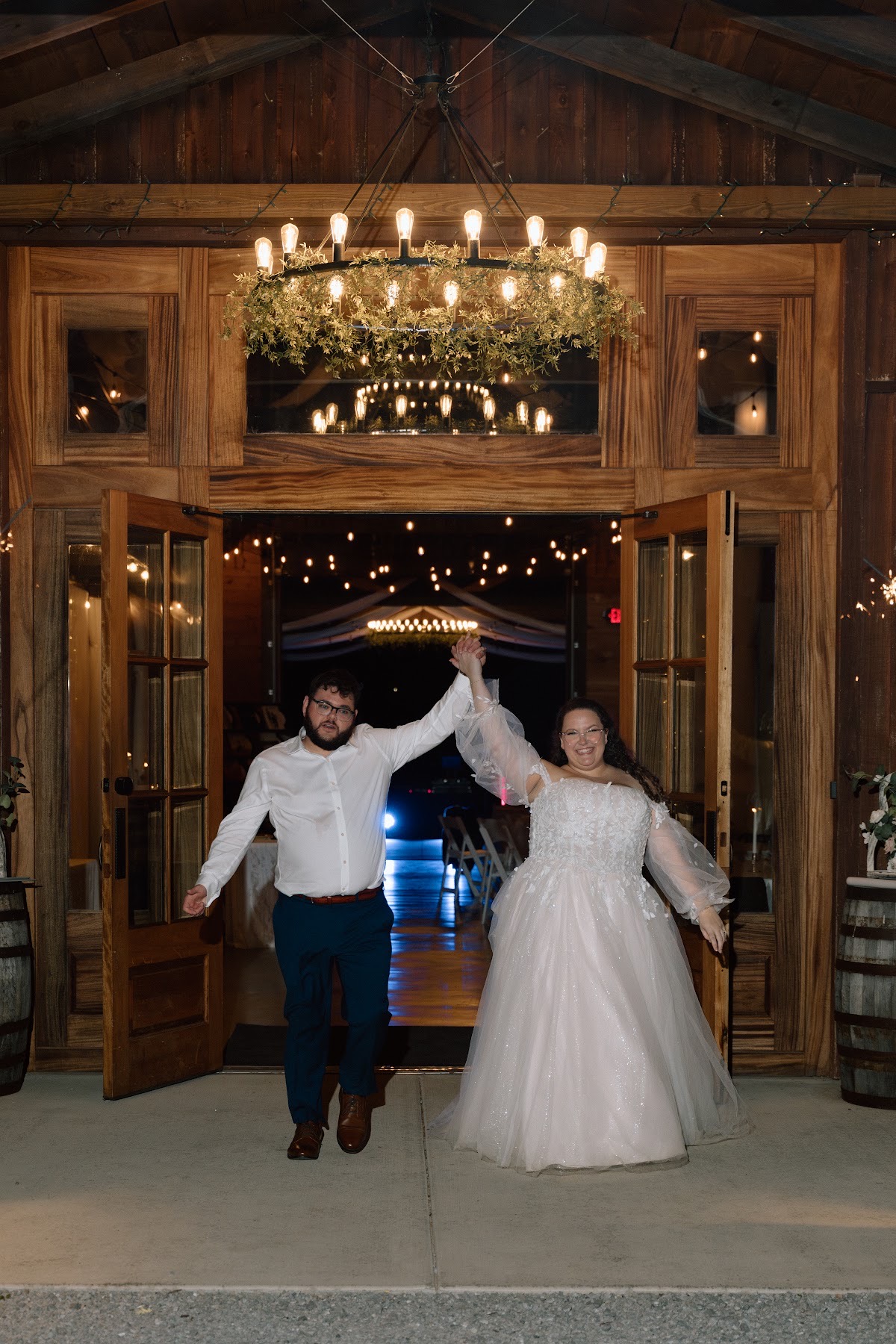 Katelynn & Justin raise their arms celebrating through open barn doors under a greenery chandelier at Covey Creek