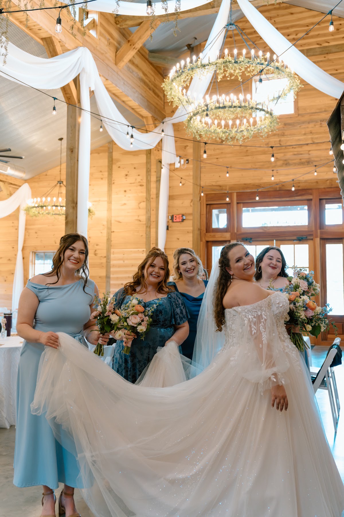 Katelynn glances back as bridesmaids lift her sparkling train under barn chandeliers at Covey Creek for Katelynn & Justin
