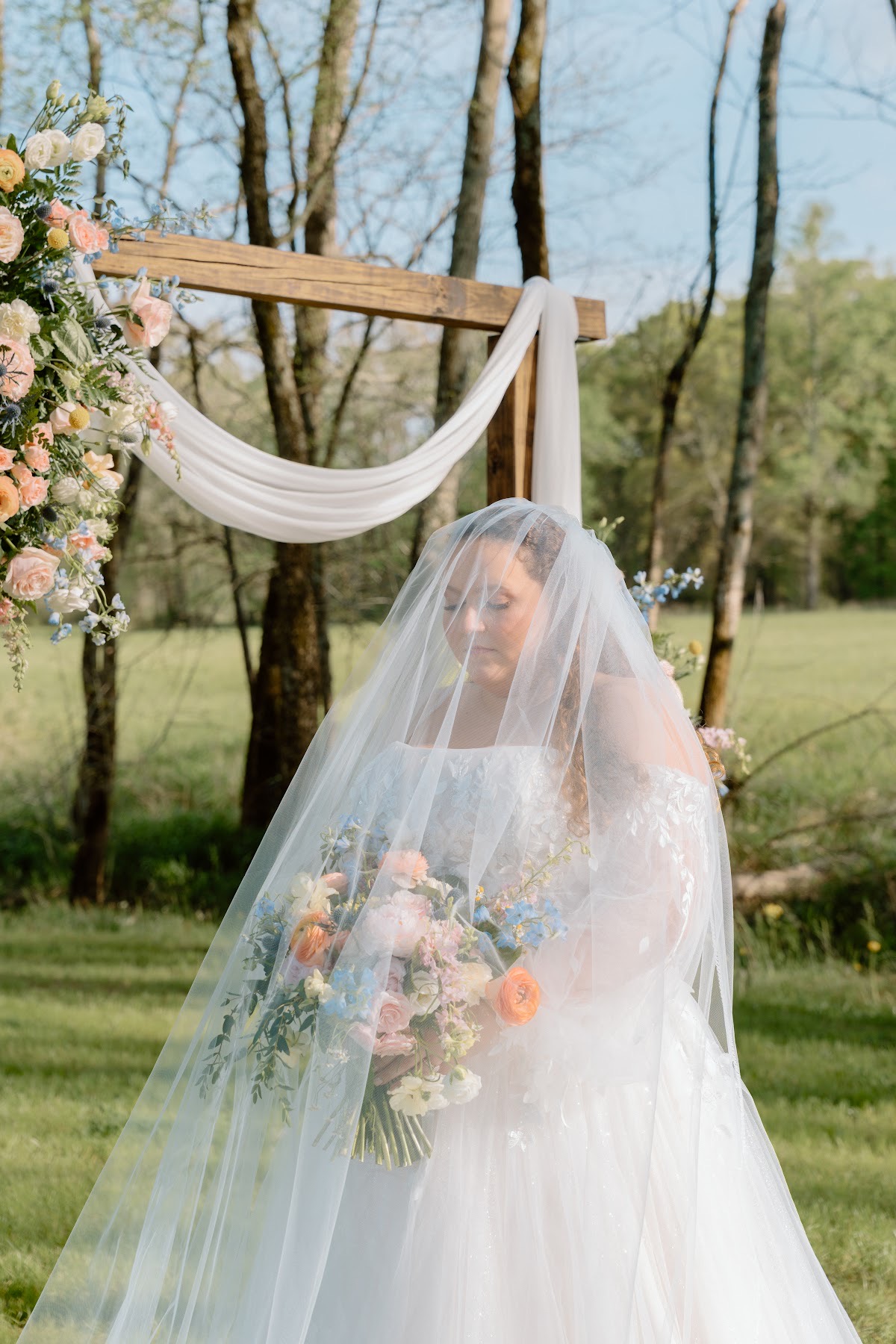 Katelynn looks down through her veil under a floral arbor, holding her bouquet at Covey Creek for Katelynn & Justin