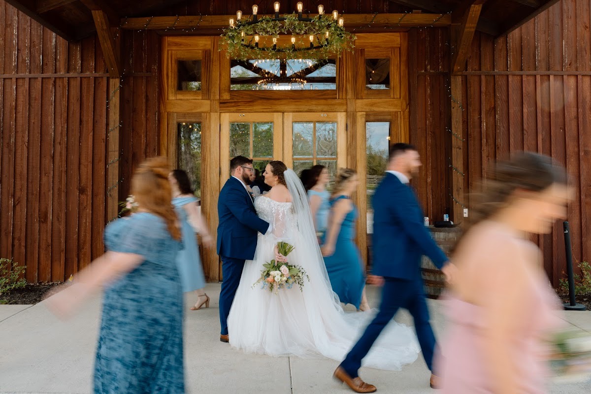 Katelynn & Justin stand still as blurred wedding party swirls past barn doors at Covey Creek