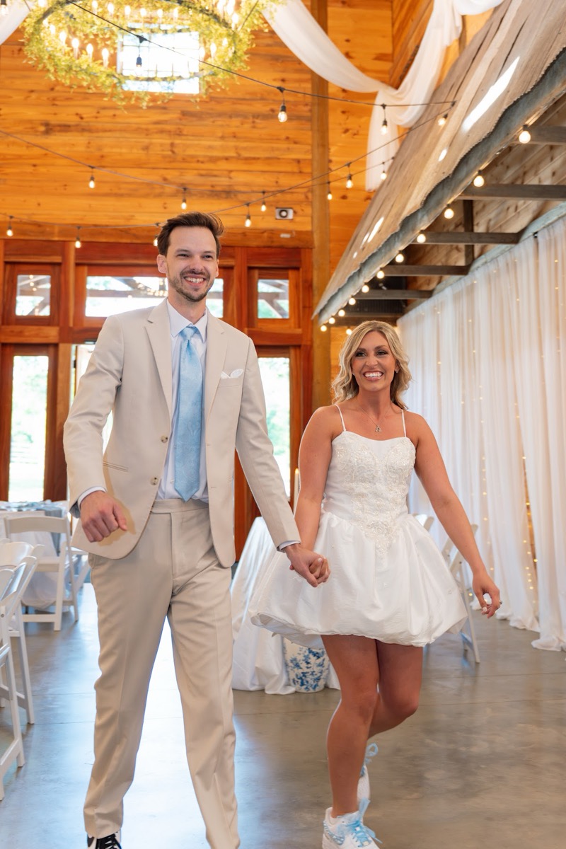 Kyley & Nick smile hand in hand in sneakers under barn drapes and string lights at Covey Creek