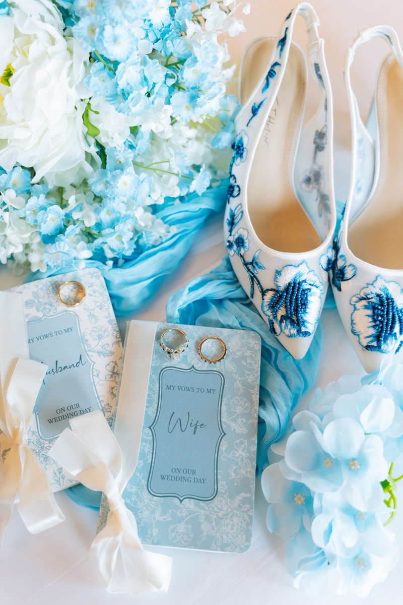 Vow books, rings, and blue floral heels arranged with blooms for Kyley & Nick at Covey Creek