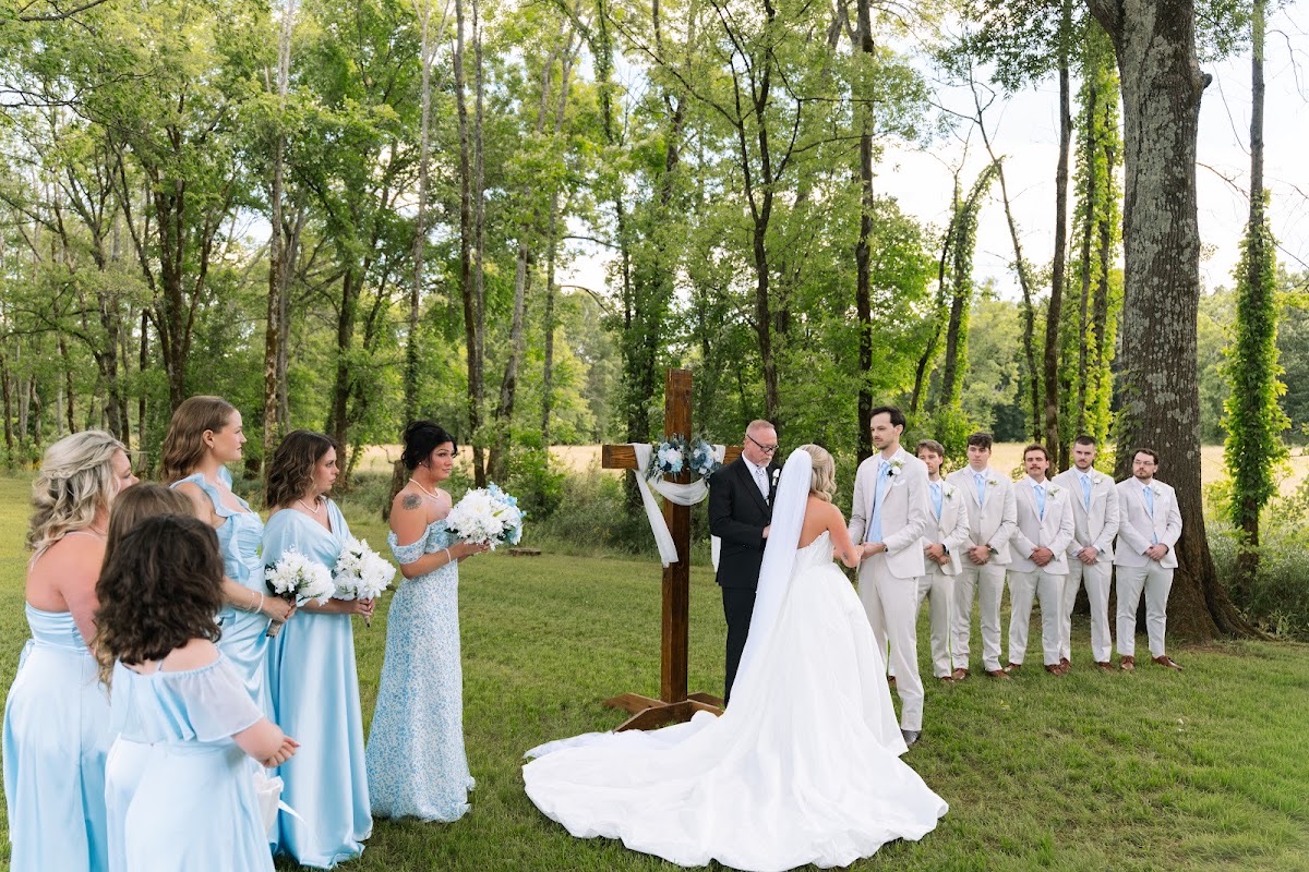 Kyley & Nick stand before a floral wooden cross with their wedding party under trees at Covey Creek