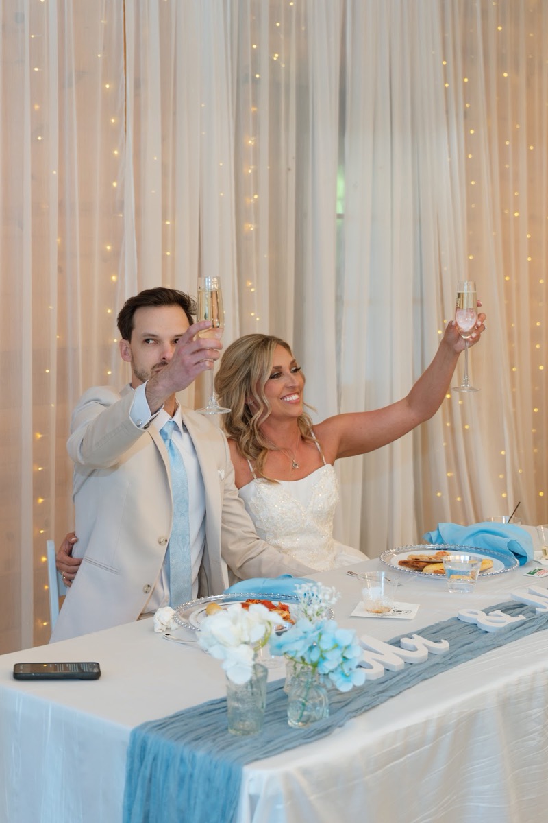 Kyley & Nick raise champagne flutes at their sweetheart table under lit drapes at Covey Creek