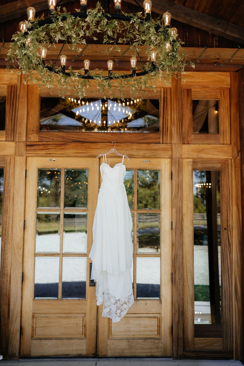 Kylie & Mason bridal gown hangs on wood glass doors beneath a greenery chandelier at Covey Creek