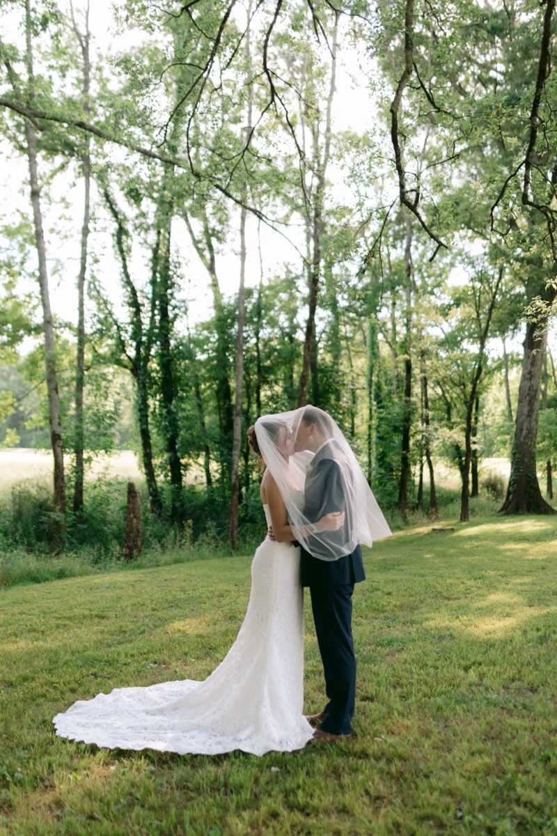 Rachel & Ben kiss under a sheer veil on sunlit grass beneath lush spring trees at Covey Creek.