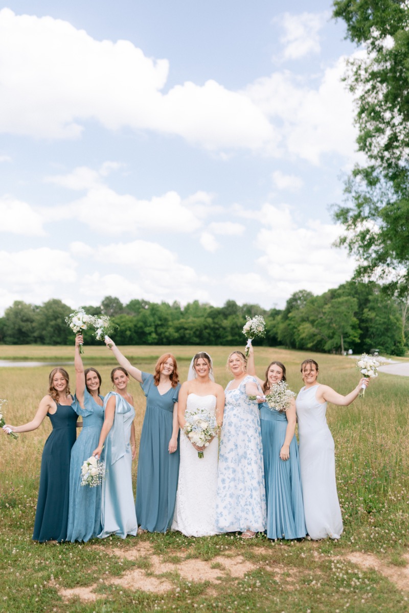 Rachel & Ben with bridesmaids raising bouquets in a sunny field by the Pond at Covey Creek.