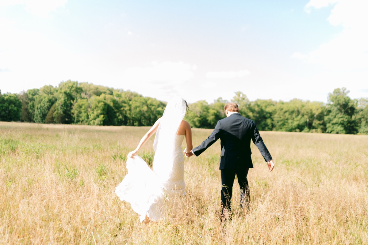 Rachel & Ben walk hand-in-hand through a sunlit golden field toward green trees at Covey Creek.