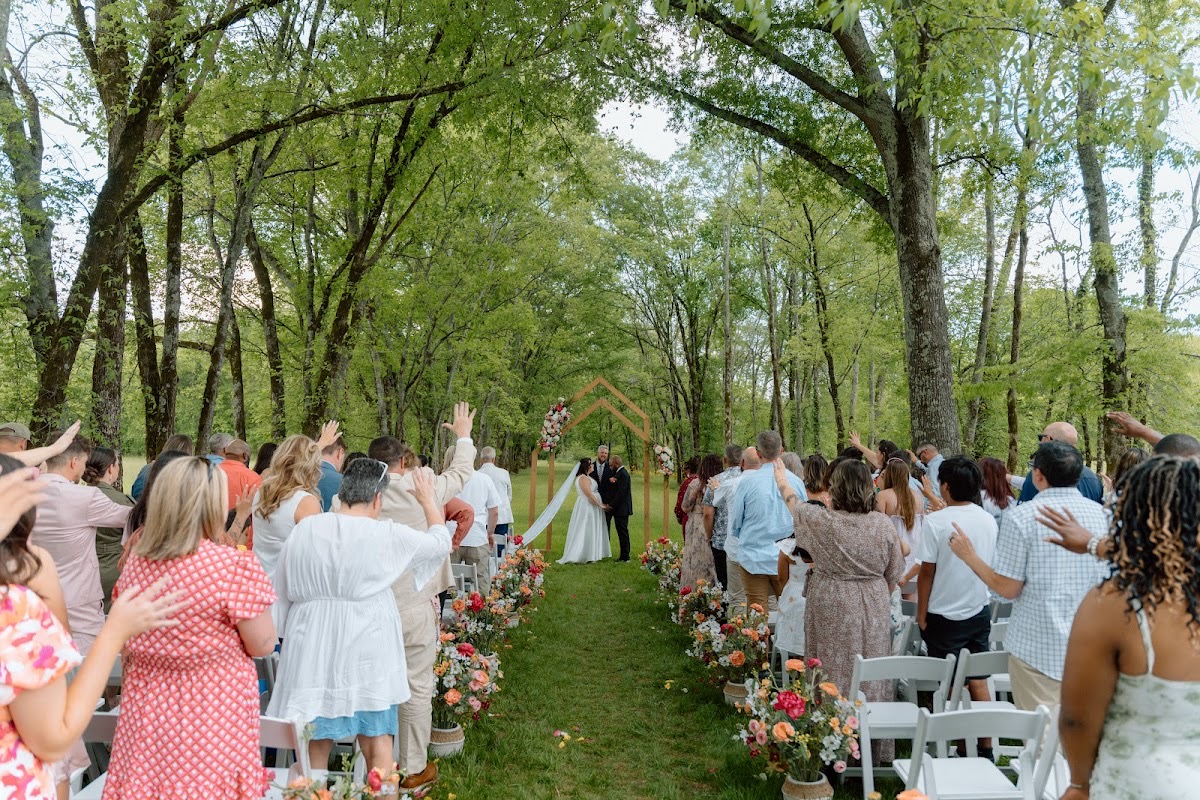 Guests raise hands toward Stephanie & Josh under the peaked wooden arch along the tree aisle at Covey Creek