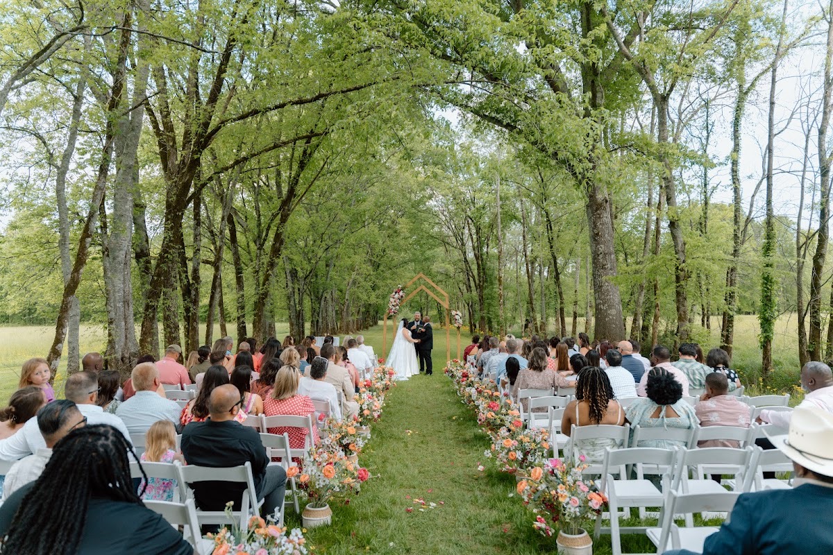 Tree canopy frames Stephanie & Josh under the wooden arch along the flower-lined aisle at Covey Creek