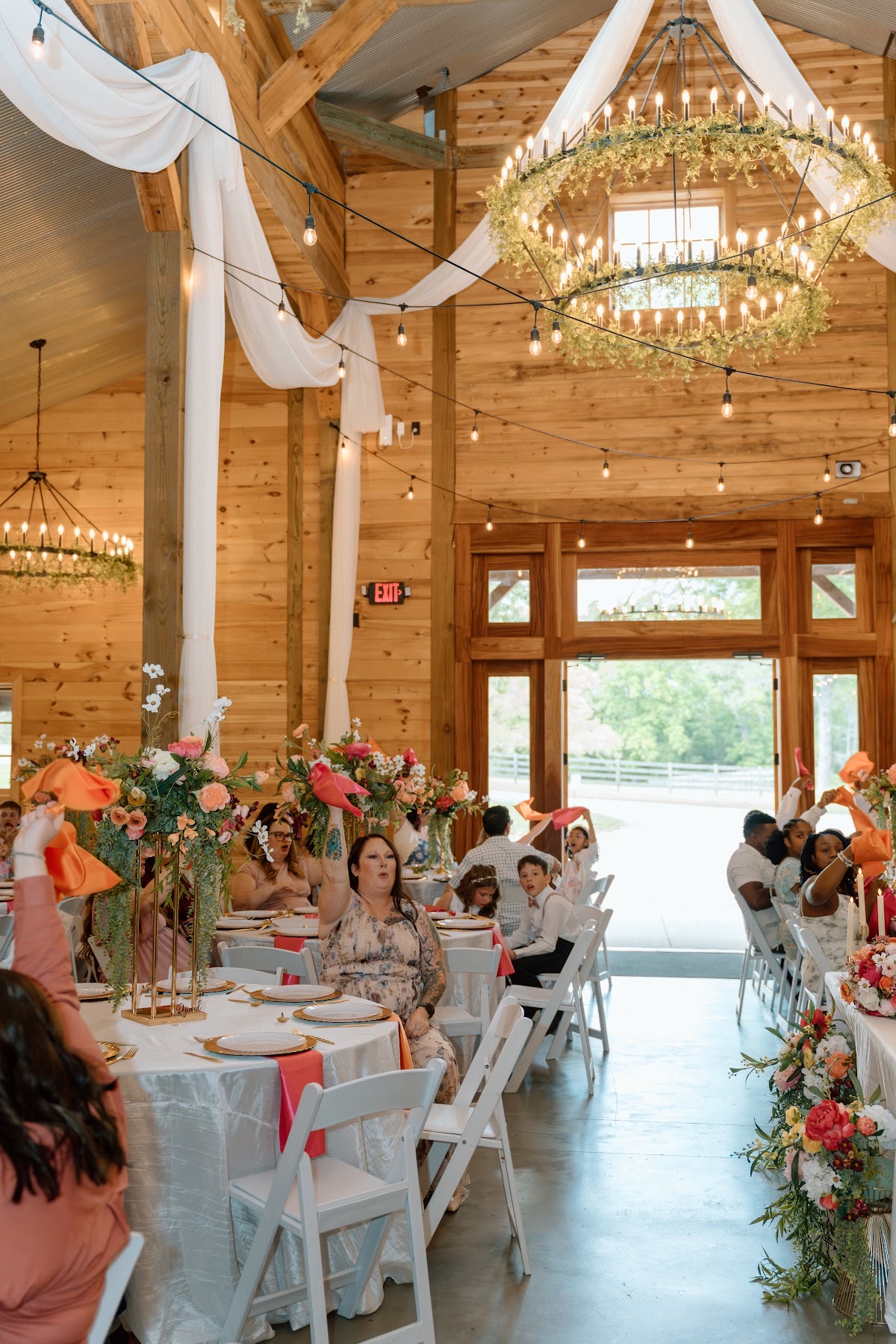 Guests raise coral napkins at round floral tables in the wood barn for Stephanie & Josh at Covey Creek