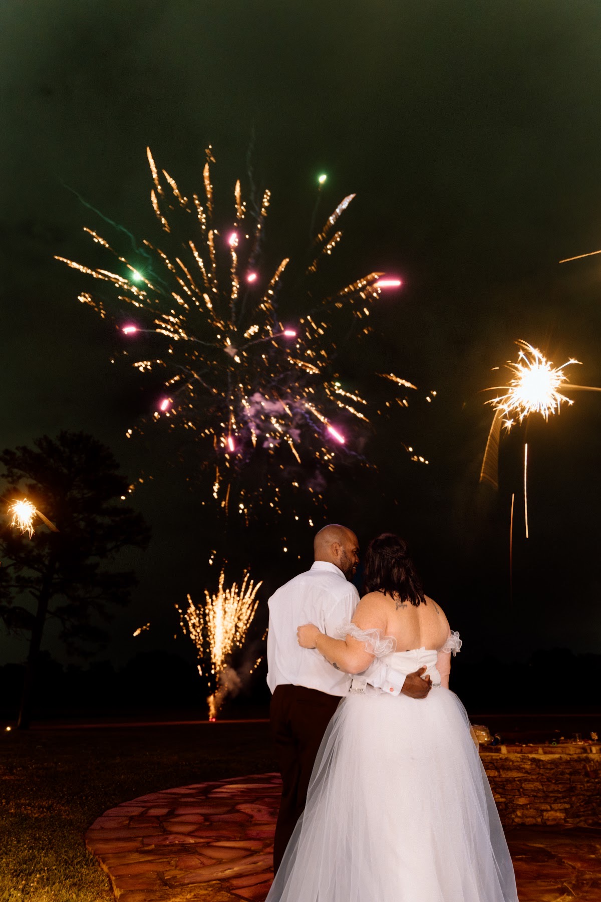 Stephanie & Josh stand arm in arm watching pink and gold fireworks burst over the field at Covey Creek