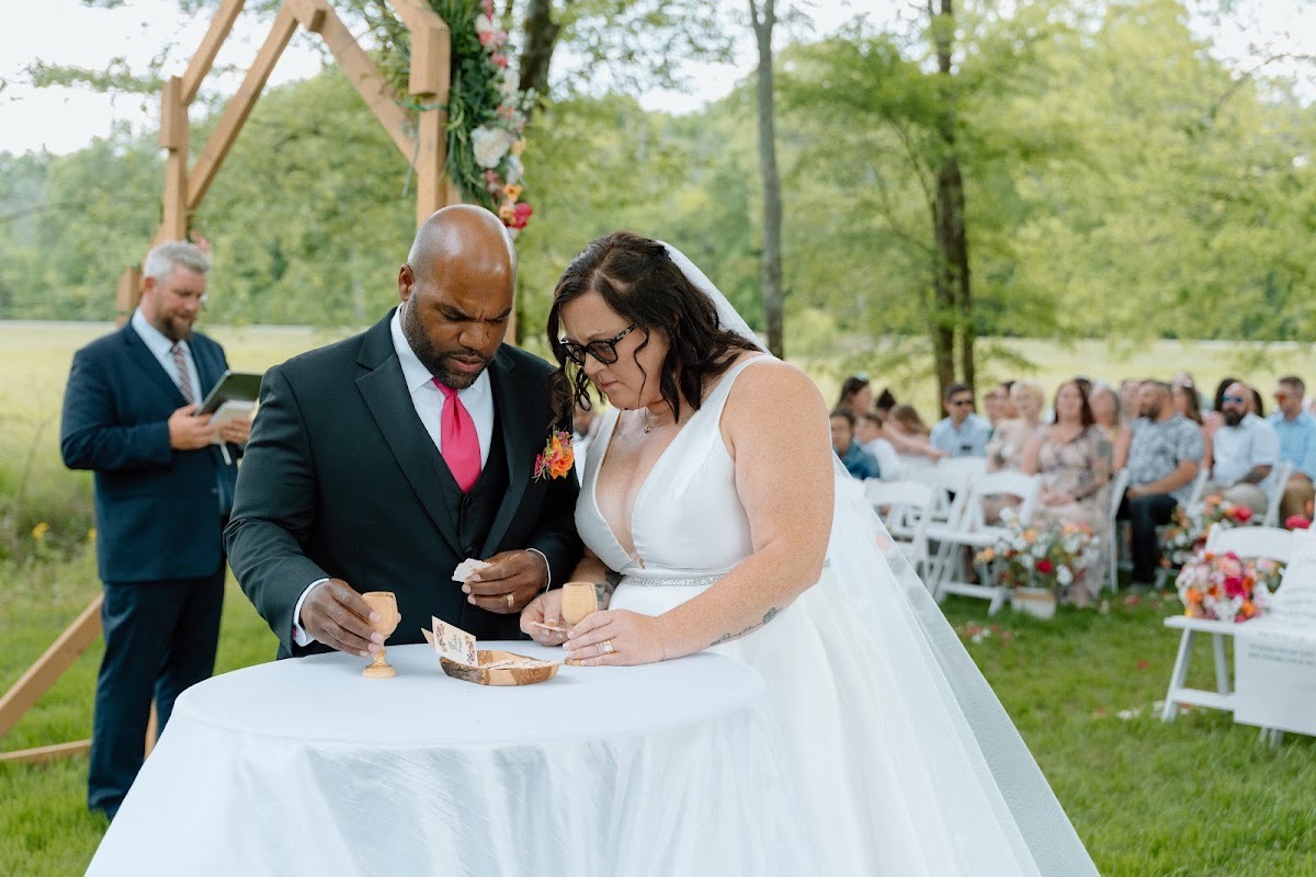 Stephanie & Josh hold wooden cups at a small table under the floral wooden arch at Covey Creek