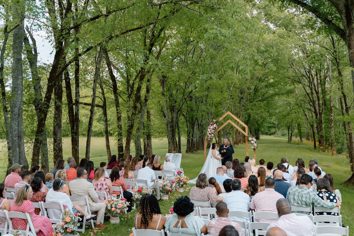Wide view of Stephanie & Josh under the peaked wooden arch before guests among the trees at Covey Creek