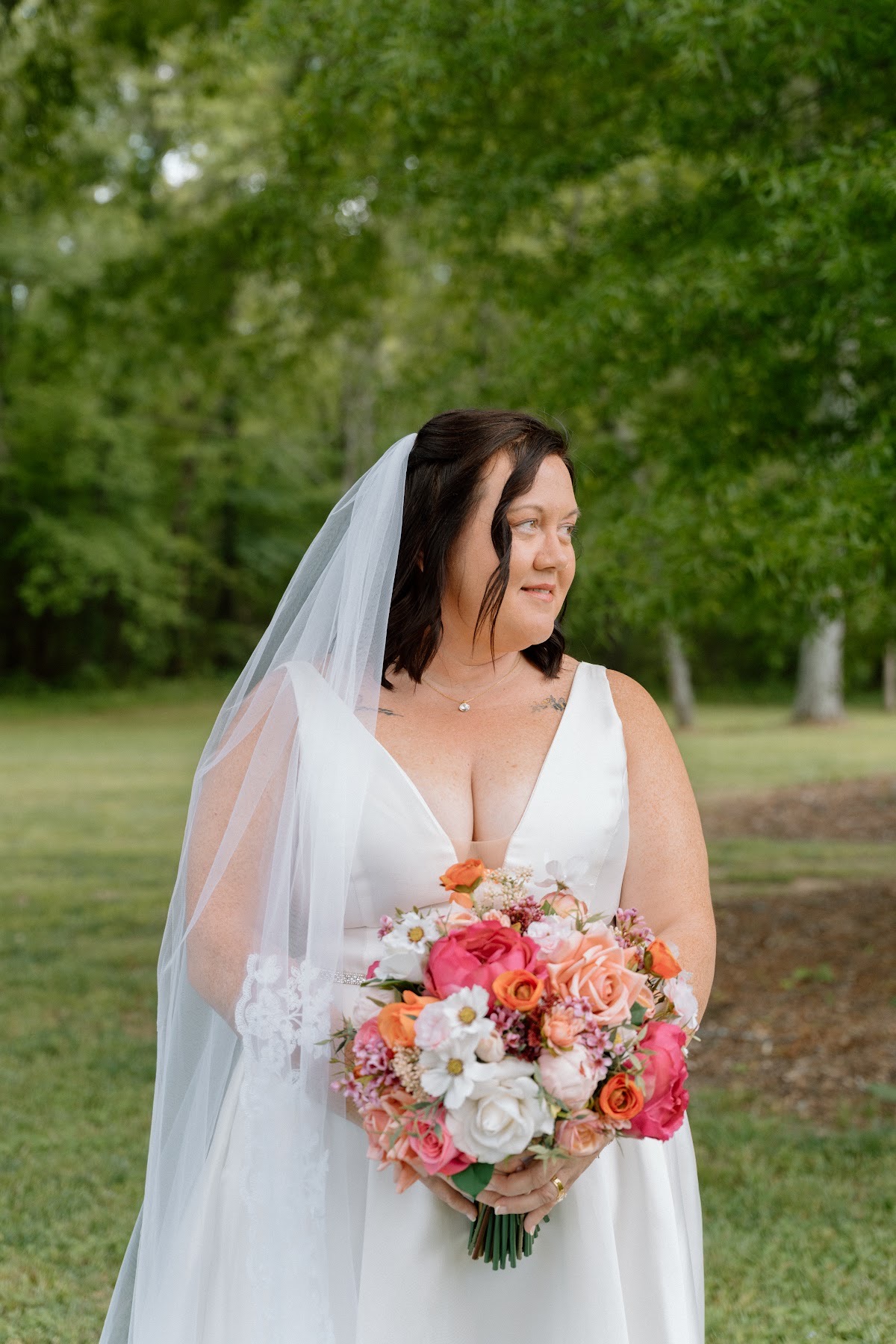 Stephanie holds her pink and orange bouquet in a lace veil against green trees for Stephanie & Josh at Covey Creek