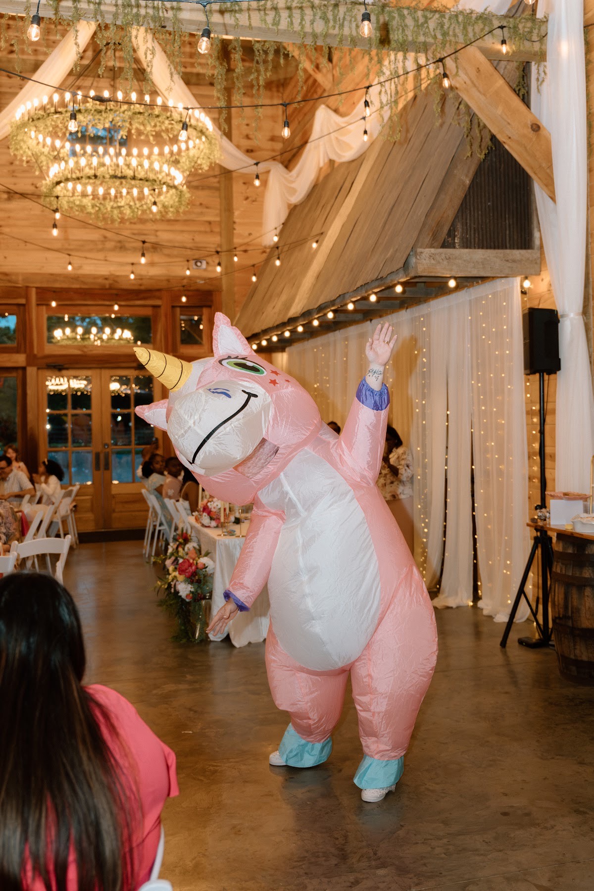 A guest in a pink inflatable unicorn dances under barn chandeliers for Stephanie & Josh at Covey Creek