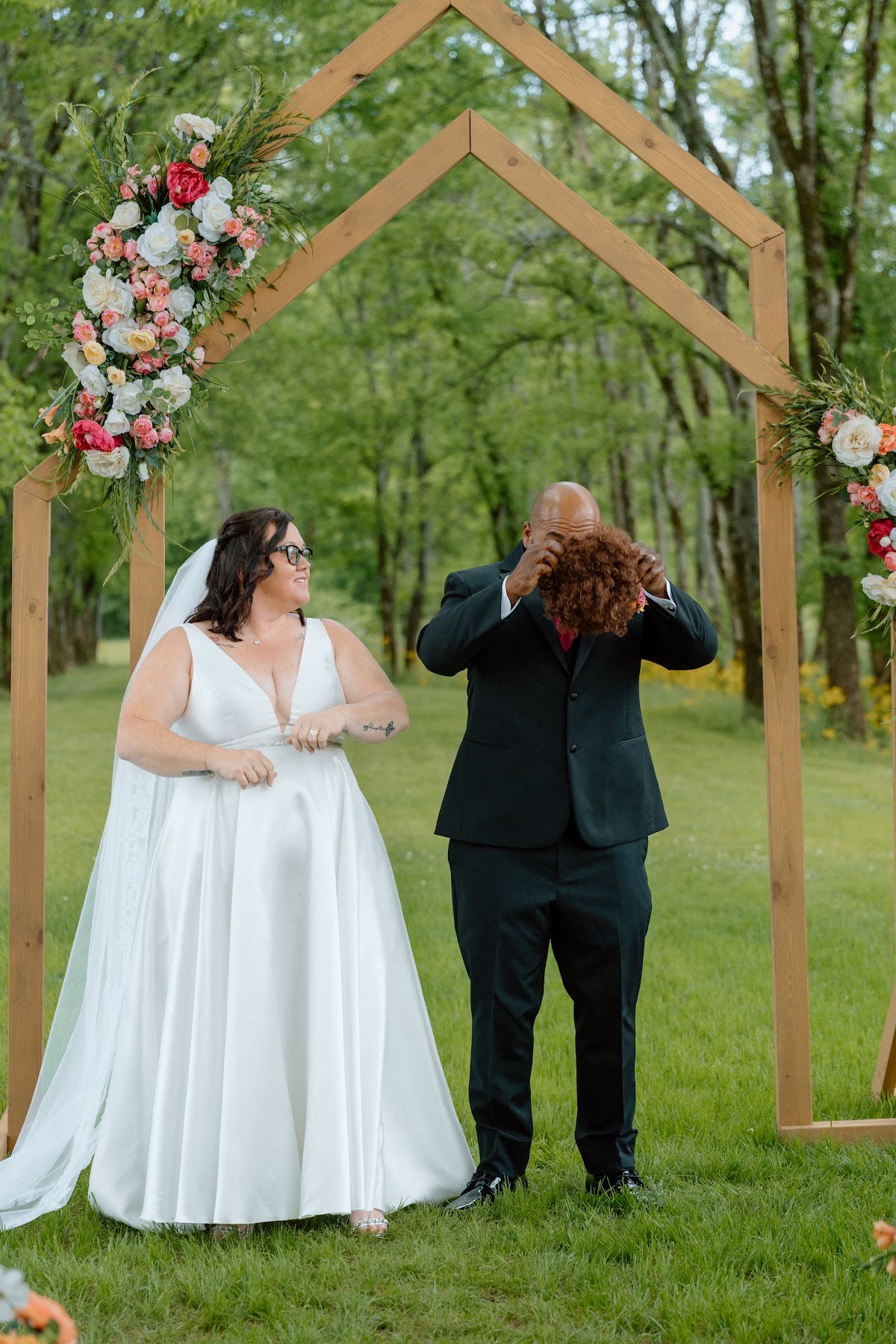 Stephanie laughs as Josh playfully hides behind her pink and orange bouquet under the arch at Covey Creek