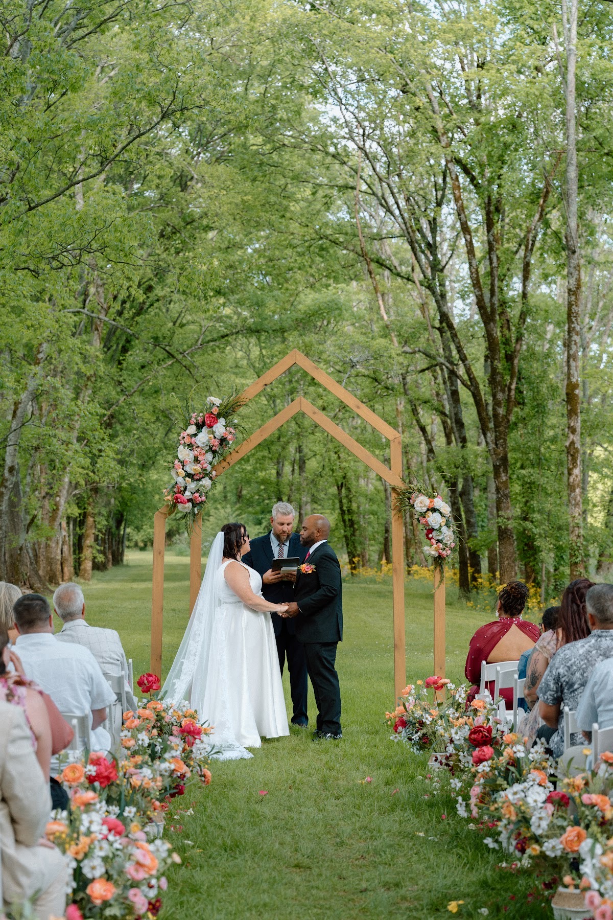 Stephanie & Josh face each other under the floral wooden arch as guests look on at Covey Creek